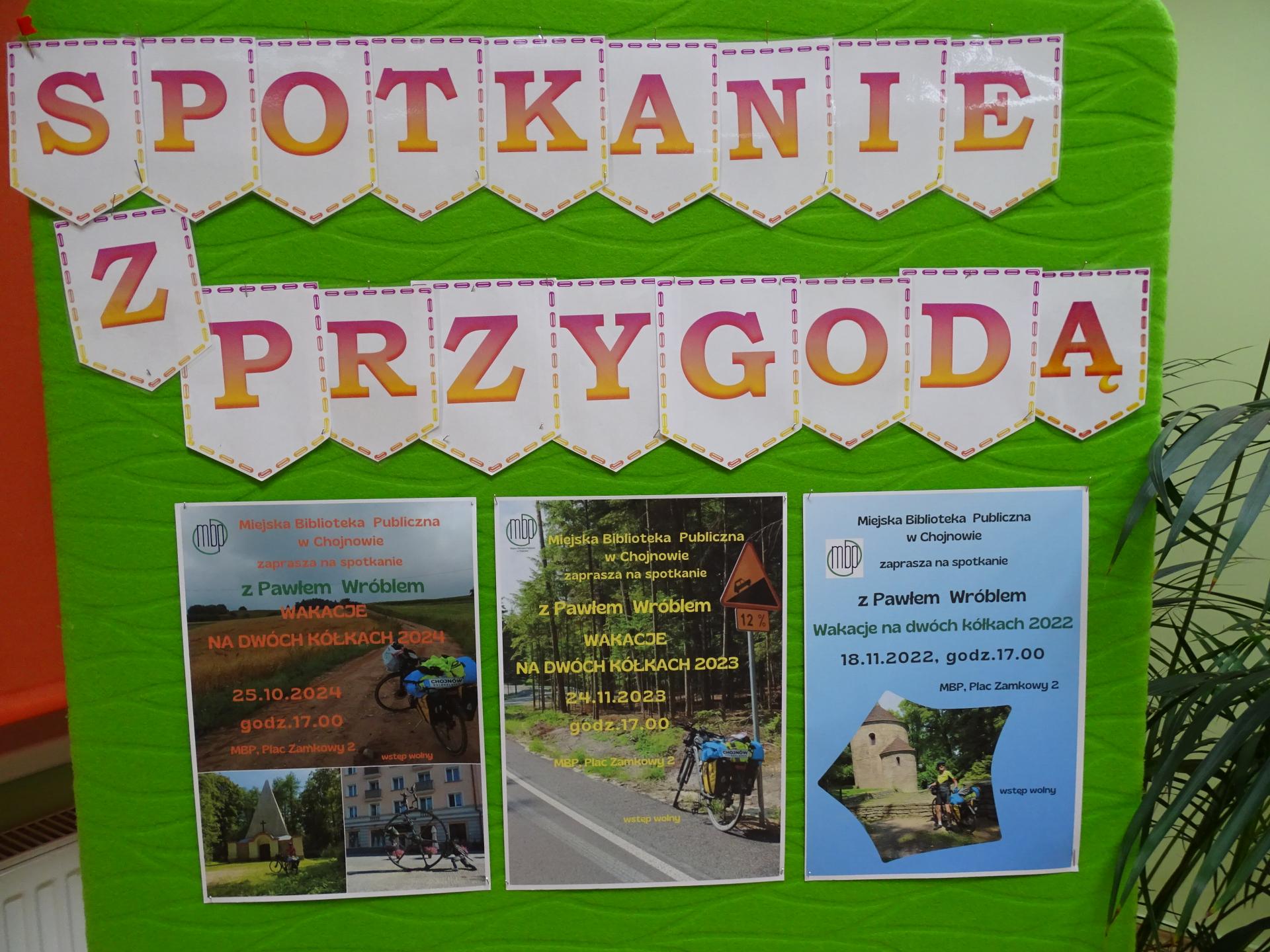 Na zielonej tablicy wisi napis Spotkanie z przygodą oraz plakaty promujące kolejne edycje spotkań z Pawłem Wróblem w latach: 2022,2023, 2024.